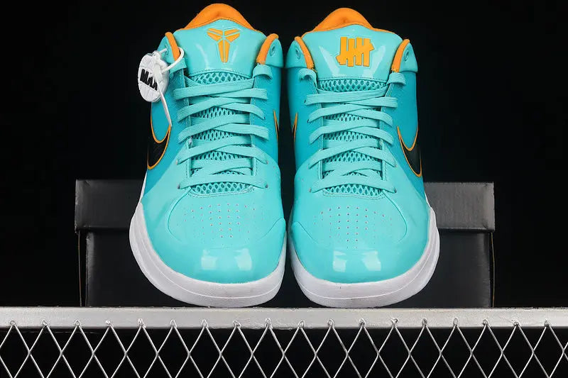 A pair of Nike Kobe 11 Low sneakers in a vibrant turquoise and orange colorway, displayed side-by-side on a black shoebox against a dark background.