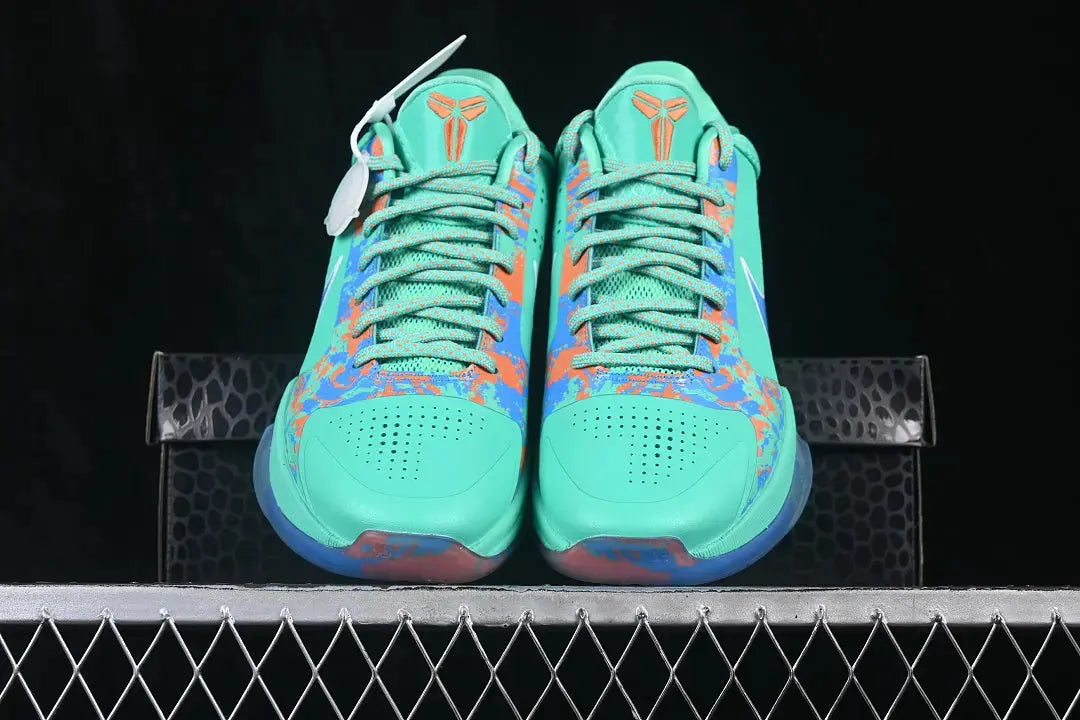 A pair of Nike basketball sneakers in a vibrant turquoise green, featuring orange and blue patterned overlays, displayed on a black textured box on a metal grid against a black background.