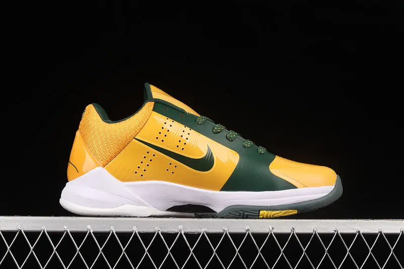 A Nike court shoe in yellow and dark green with a white midsole, shown in profile against a black background with a metal fence at the bottom.