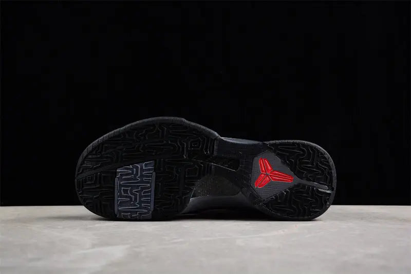 The outsole of a black sneaker with a red Tri-Ferg logo on the heel, resting on a light gray surface against a black background.