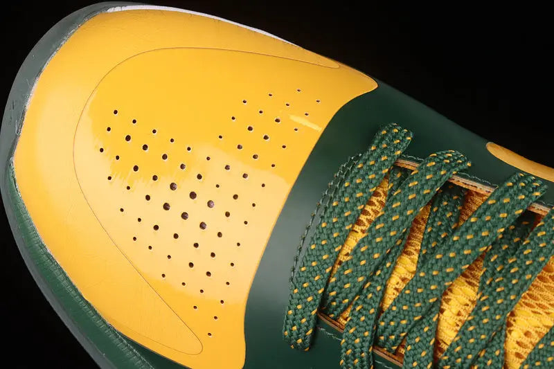 Close-up of a yellow and green sneaker showing the toe box, perforated yellow overlay, dark green side panel, and green-and-yellow braided laces against a black background.