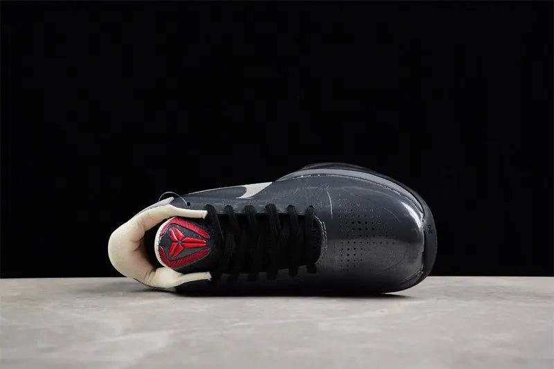 A single black and dark gray basketball sneaker shown from the side profile, resting on a light gray surface against a solid black background. The shoe features a black leather upper, black laces, a cream/off-white collar, and a red and black logo on the heel.