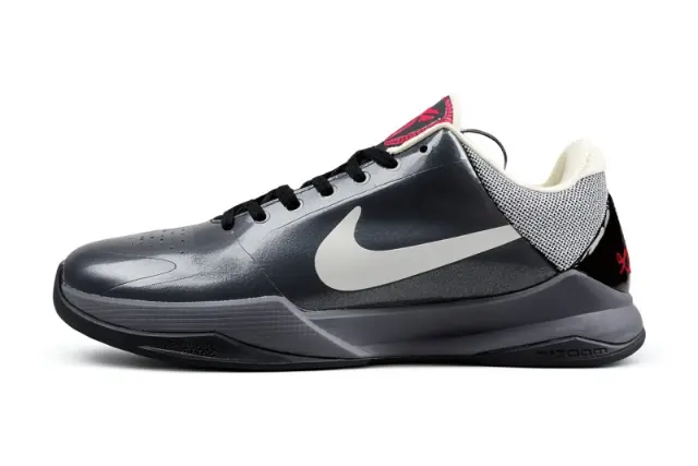 A Nike court shoe in dark gray and black with a prominent white Swoosh, black laces, a textured gray mesh collar, and a small red accent on the tongue and heel.