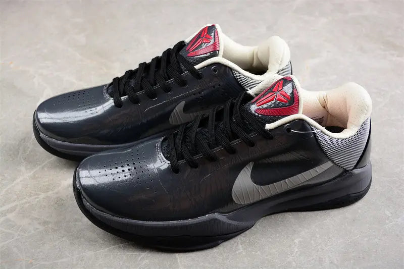A pair of Nike basketball sneakers in black and dark gray with red and white accents on the tongue and heel, displayed on a gray stone-textured surface.