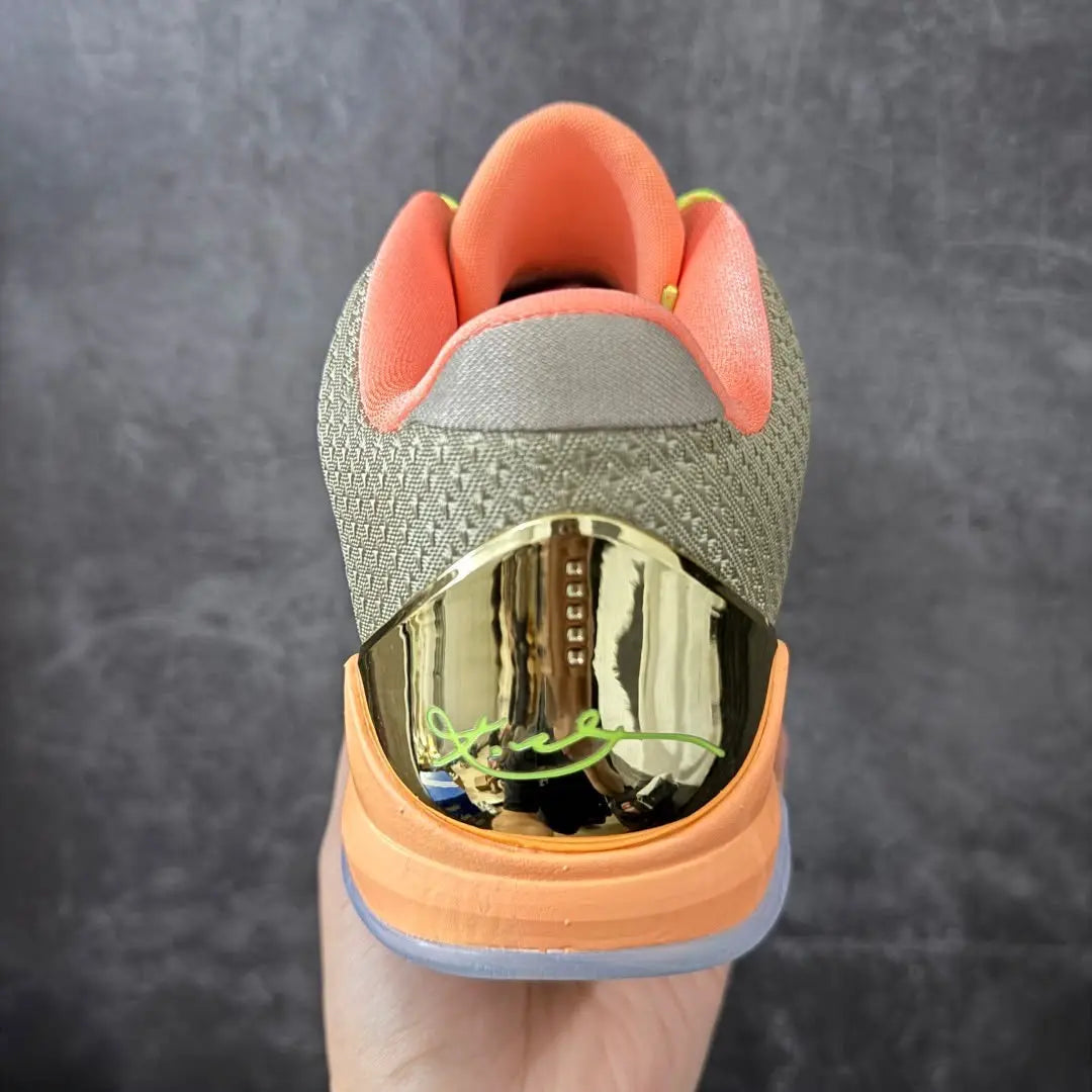 A close-up rear view of a basketball shoe held in a hand against a dark gray textured background. The shoe features a textured olive green and gray upper, a prominent metallic gold heel counter, and a bright orange midsole and lining.