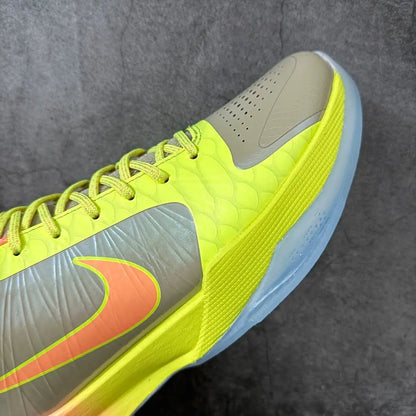 A close-up of a Nike basketball shoe in neon yellow, gray, and orange, showing the side profile, lace-up closure, textured upper, and part of the translucent blue-gray sole against a dark gray background.