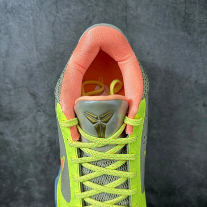 A close-up top-down view of a Nike basketball shoe, showing the neon yellow-green laces, gray mesh upper, orange lining, and a metallic gray tongue with a gold Kobe Bryant logo.