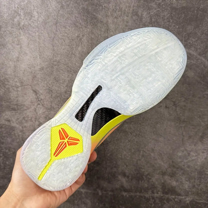 A hand holding a basketball shoe's translucent outsole, showing a yellow and red logo on the heel, a black carbon fiber pattern cutout, and a yellow and white midfoot section against a gray textured background.