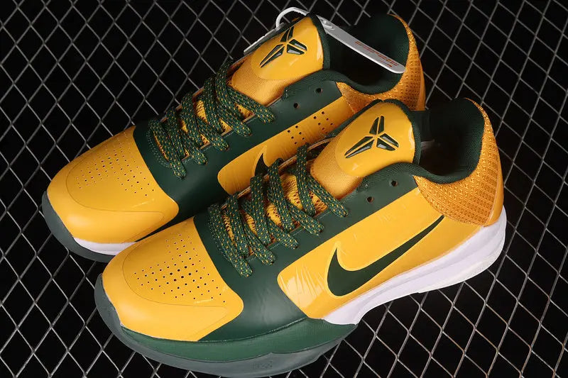 A pair of Nike basketball sneakers in yellow and green, featuring a Kobe Bryant logo on the tongue and a green Nike swoosh on the side, displayed on a black wire mesh background.