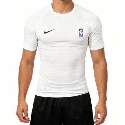 A man wearing a white Nike short-sleeve athletic compression shirt with a black swoosh on the right chest and an NBA logo on the left chest, paired with black shorts.