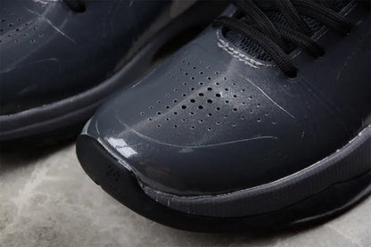 Close-up of a dark gray athletic sneaker showing the glossy synthetic leather upper, perforated toe area, black laces, and textured midsole on a stone surface.