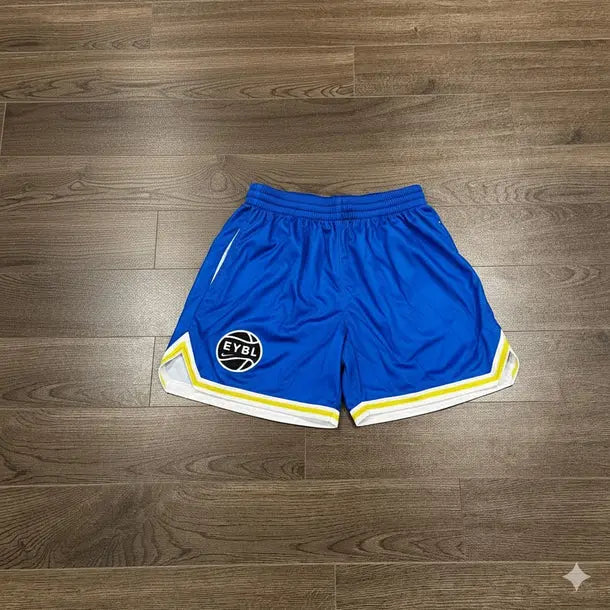 Blue athletic shorts with white and yellow stripes, featuring a circular 'EYOT' logo on the left leg, laid flat on a dark wood floor.