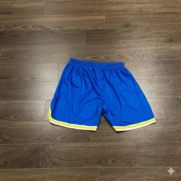Blue athletic shorts with yellow and white striped trim at the hem, laid flat on a dark wood floor.